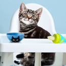 Cat sitting at a high chair with a blue bowl and green toy on a light blue background