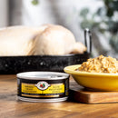 Can of shredded chicken on a wooden table with a bowl of food and roasted chicken in the background.