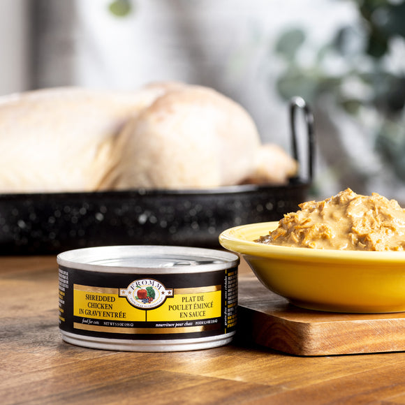Can of shredded chicken on a wooden table with a bowl of food and roasted chicken in the background.