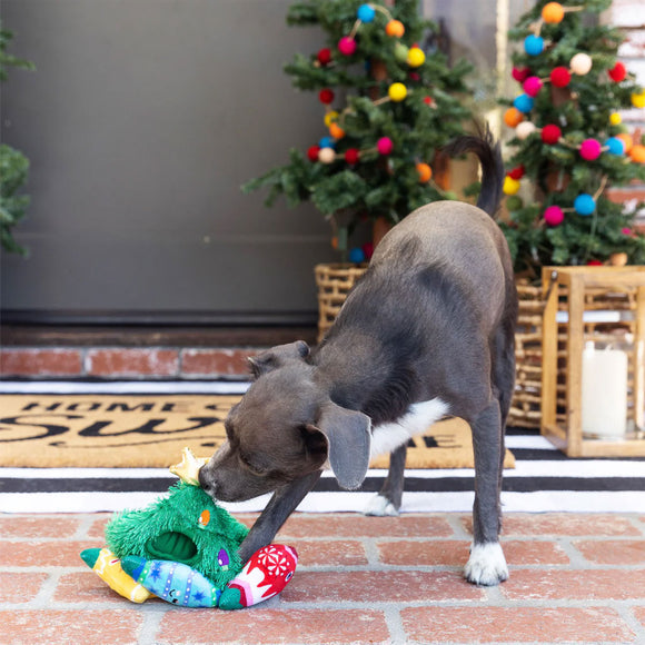 Dog playing with a Christmas-themed toy on a brick patio with decorated trees in the background.