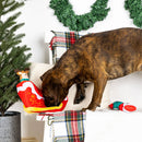 Dog playing with a Christmas-themed toy on a decorated couch with a wreath in the background.
