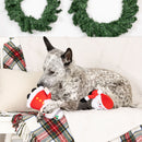 Dog sitting on a couch with Christmas decorations and a toy, surrounded by festive pillows and wreaths.