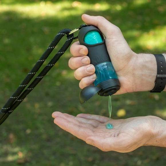 Hand holding a black and teal device with a green liquid being dispensed onto a palm against a blurred green outdoor background.