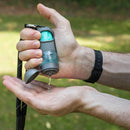Hand holding a small device with a green light, likely a dog training clicker, against a blurred natural background.