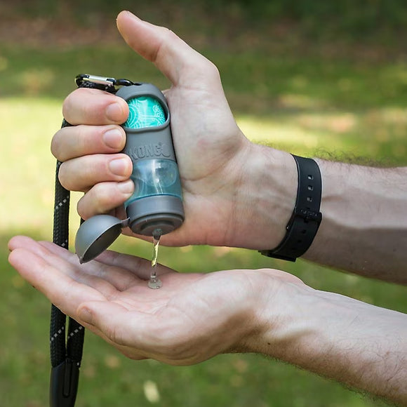 Hand holding a small device with a green light, likely a dog training clicker, against a blurred natural background.