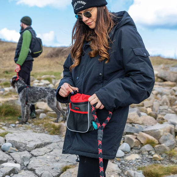 Person in a black jacket and sunglasses holding a red and black bag with a dog in the background on a rocky landscape.