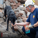 Man in blue shirt and hard hat with a black dog amidst rubble, holding Nulo jerky.