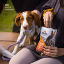 Dog sitting next to a person holding a Nulo Freestyle product package outdoors.