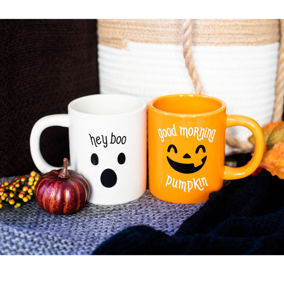 Two mugs, one white with a ghost design and 'hey boo' text, the other orange with a pumpkin face and 'good morning pumpkin' text, on a textured surface with pumpkins and leaves.