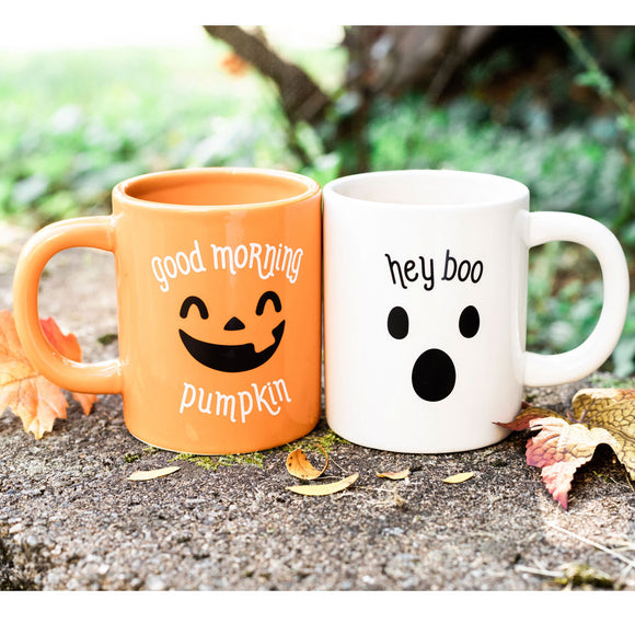 Two Halloween-themed mugs, one orange with a pumpkin face and 'Good morning pumpkin' text, the other white with a ghost face and 'hey boo' text, on a natural background.