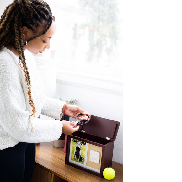 Person opening a pet memory box with a photo of a dog and a tennis ball on a table.