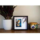Framed picture of a cat with a bracelet on a table next to a plant and decorative pot.