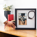 Framed picture of a dog with a black frame on a wooden surface, with a hand reaching towards it.