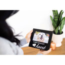 Person holding a black photo frame with a dog picture and 'Forever in my heart' text, plant and candle in the background.