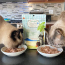 Two cats eating from bowls with a package of Coochie's Hairball Plus cat food in the kitchen.