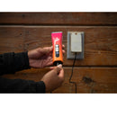 Hand holding a pink and black electronic device near a wall socket on a wooden background