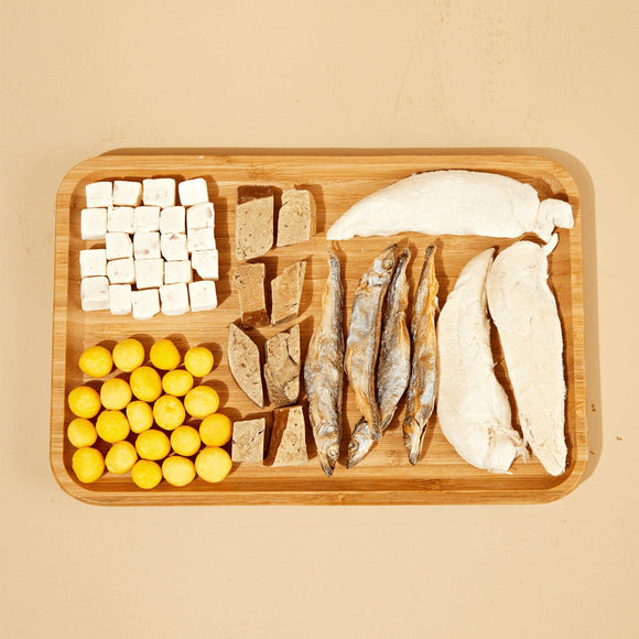 Wooden cutting board with various ingredients including fish, tofu, and eggs on a beige background