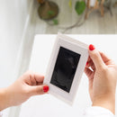 Person holding a white photo frame with a black photo inside, on a blurred background.