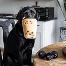Black dog holding a plush toy resembling a cup with a face in its mouth, with more similar toys on a table.