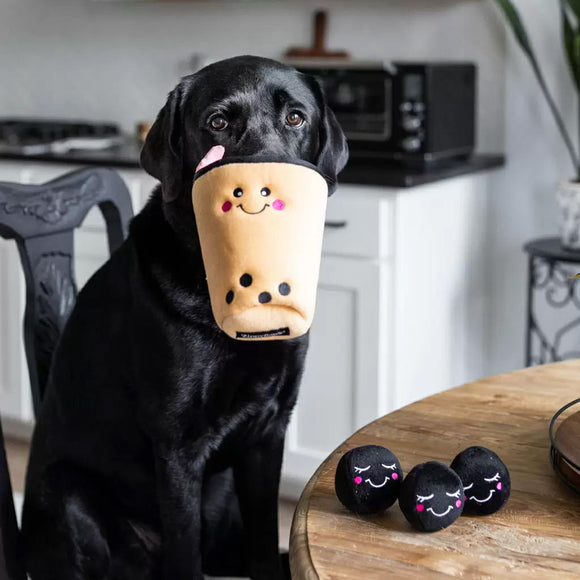 Black dog holding a plush toy resembling a cup with a face in its mouth, with more similar toys on a table.