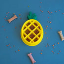 Yellow pineapple-shaped pet feeder filled with kibble on a blue background with scattered dog treats.