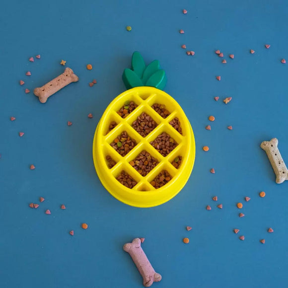 Yellow pineapple-shaped pet feeder filled with kibble on a blue background with scattered dog treats.